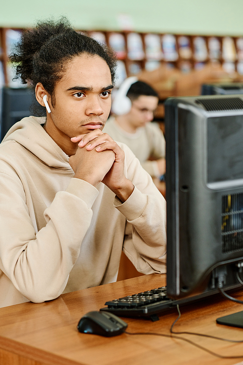 Young Black student wearing wireless earphones sitting in university library looking at computer screen thinking about task