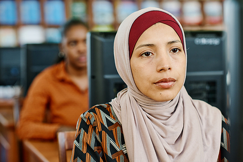 Portrait of young Muslim woman wearing hijab sitting in university library looking at computer screen