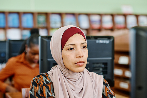Portrait of young Muslim woman wearing hijab sitting in university library working on desktop computer
