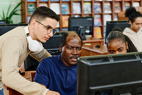 Young Middle Eastern man and Black woman helping mature man to find something in Internet in university library