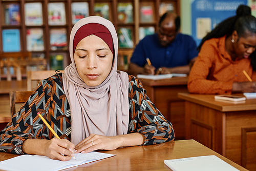 Young adult immigrant Muslim woman wearing hijab sitting at desk in classroom making notes during lesson
