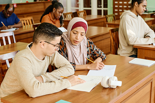 Middle Eastern man and woman in hijab sitting at table in classroom working on difficult task together