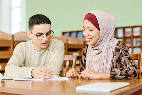 Young man and woman in hijab sitting at desk in classroom having lesson for immigrants working on writing task together