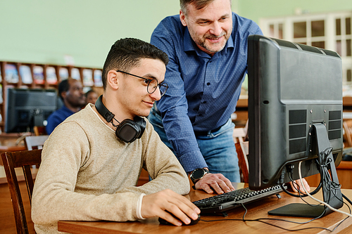 Cheerful mature teacher watching task his student finishing doing using desktop computer in library