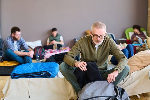 Retired man putting folded casualwear into bag while sitting on couchette against young male and female refugees resting on their beds