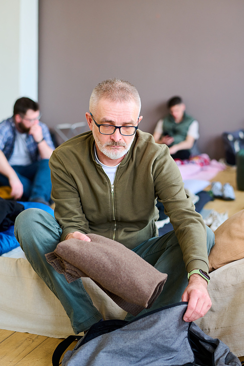 Mature man in eyeglasses and casualwear packing bag while sitting on bed or couchette in refugee camp for people in need and trouble