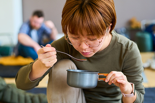 Young Chinese female refugee eating soup or other self prepared food in refugee camp against other temporarily homeless people