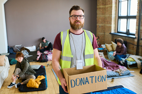 Bearded male volunteer with donation box looking at camera in refugee camp while standing among homeless people resting on beds