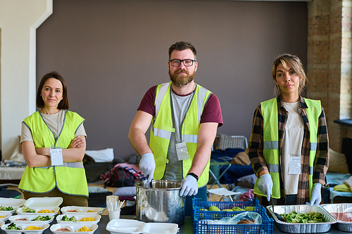 Contemporary volunteers in uniform and gloves standing by table with food prepared for refugees, homeless people or those in need