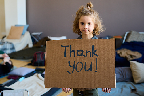 Cute blond little boy with cardboard poster saying thank you looking at camera while standing against sleeping places in refugee camp