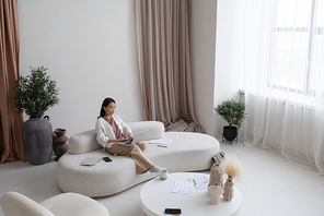 Young Asian female employee or freelancer with laptop sitting on sofa in large living room of modern apartment while networking