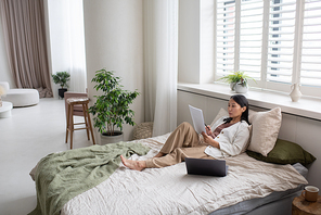 Young confident Asian female accountant in casual clothes sitting on bed and looking through financial papers while working from home