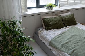 Part of comfortable bedroom with double bed standing by windowsill with domestic plants in flowerpots in large contemporary apartment