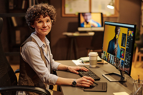 Portrait of young female designer smiling at camera while using tablet pen for editing photos on computer screen at office