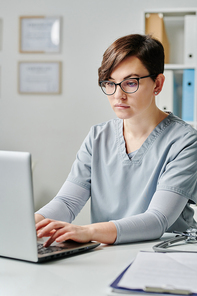 Young doctor in eyeglasses and medical uniform consulting patients online whil.e siting by desk in front of laptop in medical office