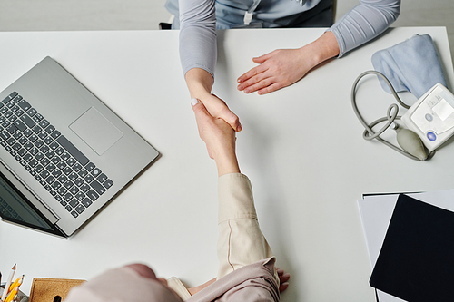 Above angle of handshake of Muslim female patient and young clinician welcoming one another over desk with laptop and tonometer