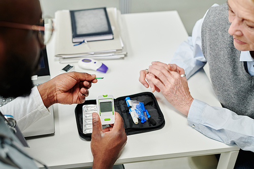 Hands of young African American doctor showing mature patient glucometer before medical test of sugar level in blood