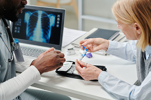 Mature female patient showing glucometer over open medical kit while sitting by desk in front of African American male doctor in hospital