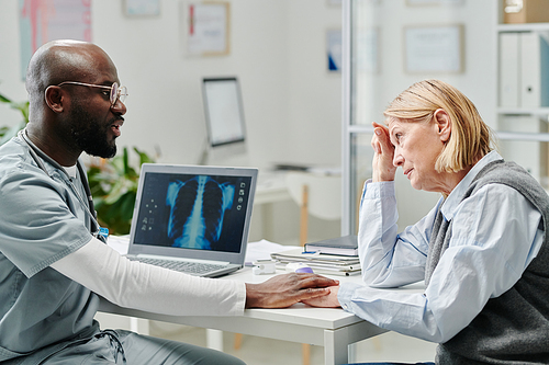 Young doctor comforting mature female patient with dangerous disease while sitting by workplace in front of her and covering her hand