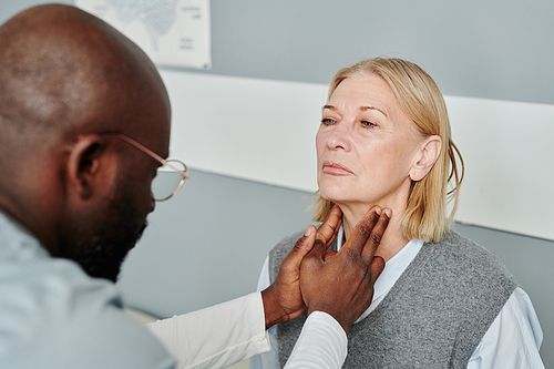 Mature blond female patient having checkup of her thyroid glands by African American male endocrinologist in medical office