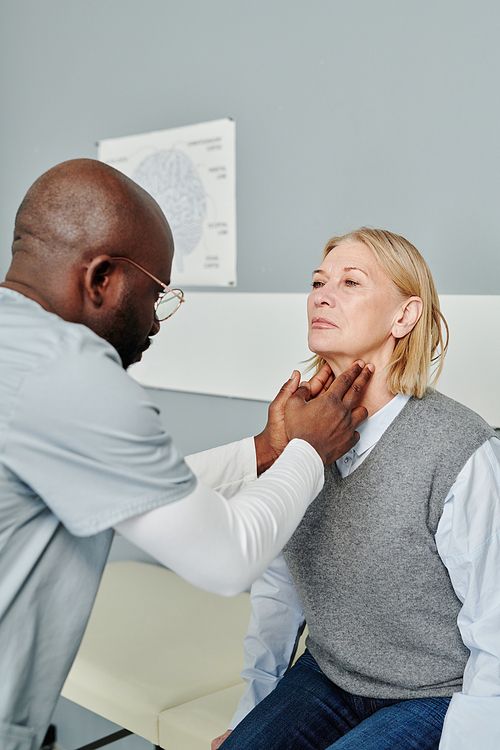 African American male endocrinologist checking thyroid of mature female patient in casualwear sitting by wall in front of him in hospital