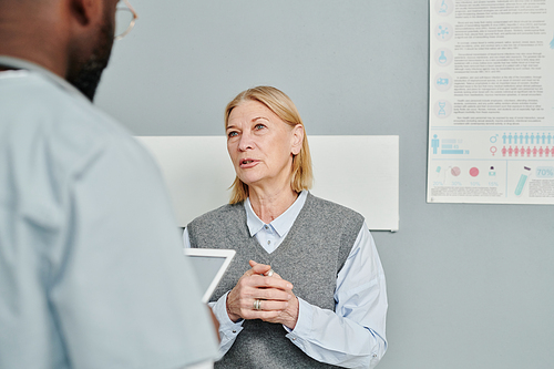 Contemporary mature female patient in casualwear describing her symptoms to doctor while sitting in front of him in hospital