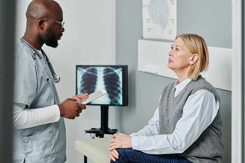 Mature female in casualwear discussing her diagnose with confident African American male doctor in uniform standing in front of her