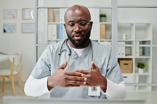 Young confident physician in eyeglasses and uniform consulting online patient while sitting by workplace in front of computer in office