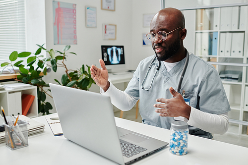 Young confident African American online doctor in uniform sitting by desk in front of laptop in clinics and giving medical advice to patient