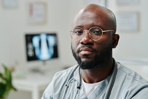 Young serious African American male general practitioner in blue uniform and eyeglasses sitting by workplace in front of camera in hospital