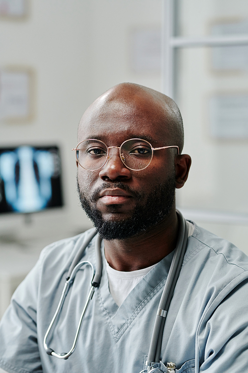 Young confident black man in uniform and eyeglasses looking at camera while sitting by workplace against computer with x-ray on screen