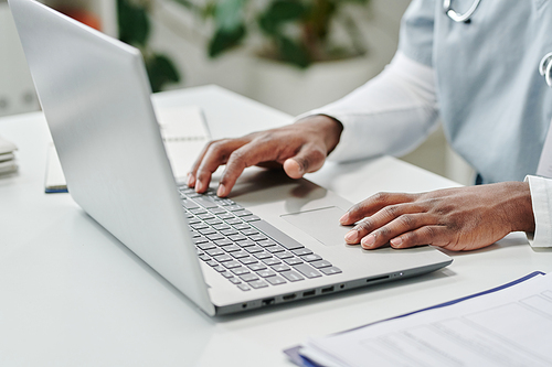 Hands of young contemporary African American male online doctor over laptop keyboard answering questions of patients in clinics