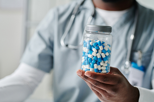 Hand of young African American male physician holding plastic jar of pills while showing it to patient during online consultation in clinics