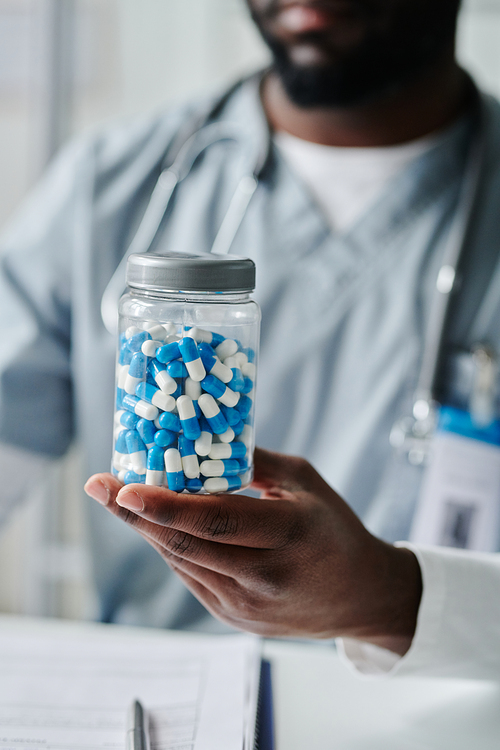 Big transparent plastic jar with pills held by contemporary male clinician in blue uniform sitting in front of camera in medical office