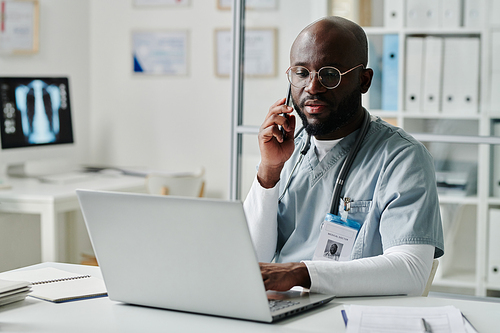 Young confident physician in medical scrubs and eyeglasses communicating with patients online and on mobile phone