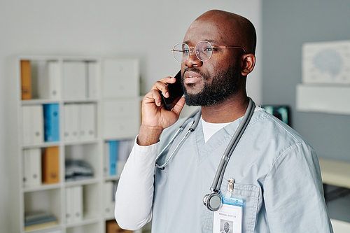 Confident male clinician with stethoscope on neck talking to patient on mobile phone while standing in front of camera in medical office