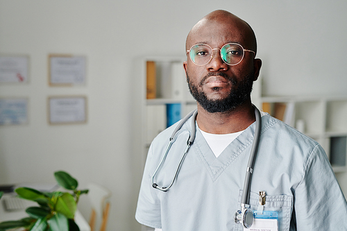 Young contemporary African American male clinician in eyeglasses and blue uniform standing in medical office and looking at camera