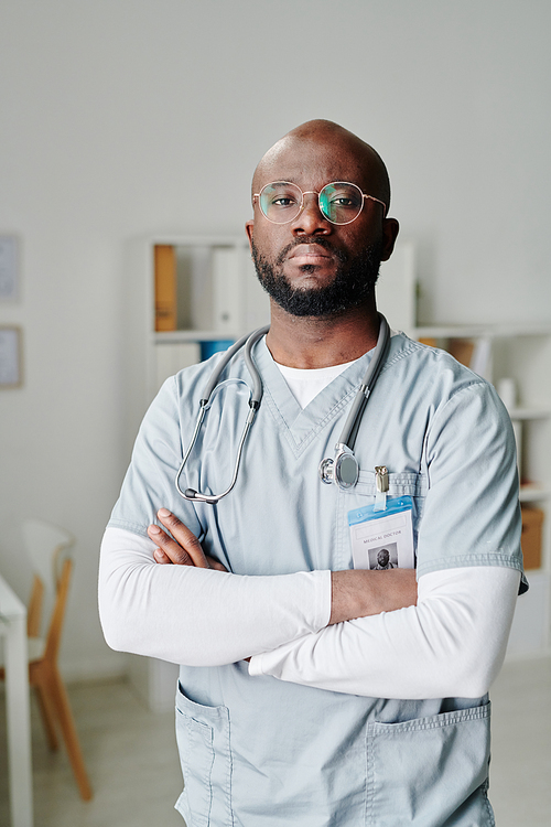 Serious young African American male clinician with phonendoscope on his neck crossing arms by chest while standing in medical office