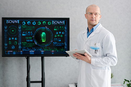 Serious mature doctor in white coat standing near the big display and using tablet pc during conference