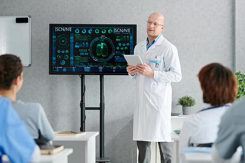 Mature lecturer in white coat standing at big display with medical charts on it and giving lecture to students