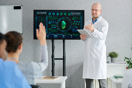 Mature doctor in lab coat with tablet pc asking question and pointing at girl who raising her hand at desk during seminar