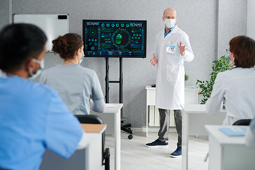 Professor in white coat and mask giving lecture about medical innovation for interns at desks at class