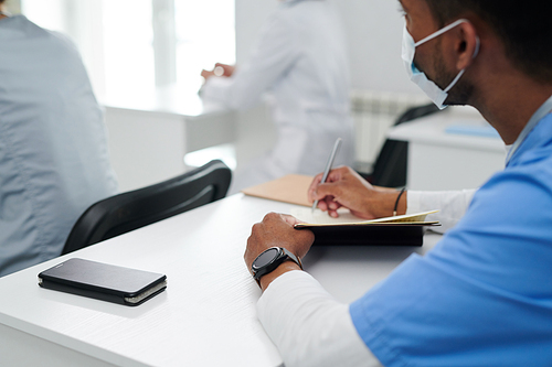 Close-up of medical student in mask sitting at desk during training and making notes in notebook