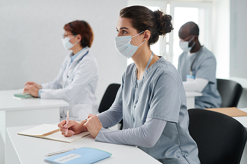 Young woman in mask sitting at desk with notebook and listening to speaker at medical training together with her colleague in background