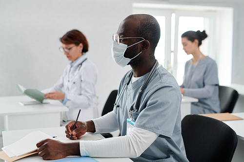 African young doctor in uniform and mask making notes in notebook at desk during medical training at class