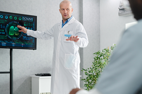 Mature doctor in white coat pointing at digital display with charts and talking to students during training