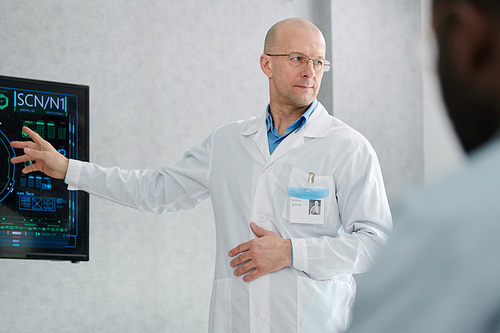 Mature speaker in white coat pointing at digital display during his lecture for medical students