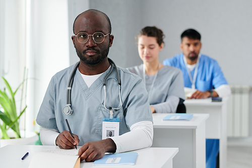 Young African doctor in eyeglasses making notes while sitting at desk during studying at medical college