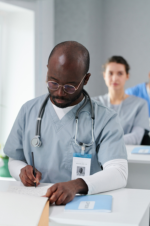 African medical specialist in uniform sitting at a lecture on professional development and making notes in notebook