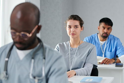 Young woman in medical uniform sitting at desk and listening to speaker during medical training